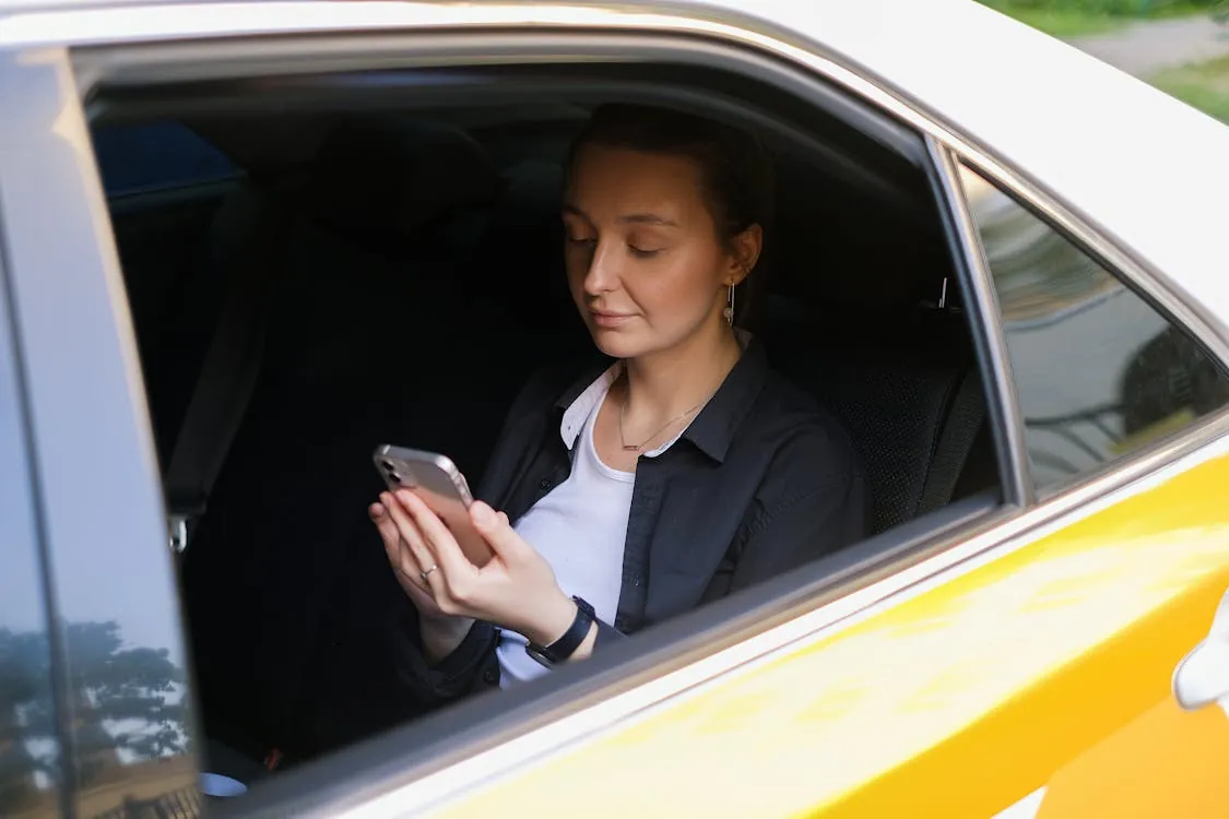 The photo shows a young woman sitting inside a yellow car, engaged with her smartphone, with a relaxed and content expression on her face, suggesting a moment of calm or leisure during a drive or break. It would suit an article about urban commuting, travel relaxation, or the use of technology during road trips.