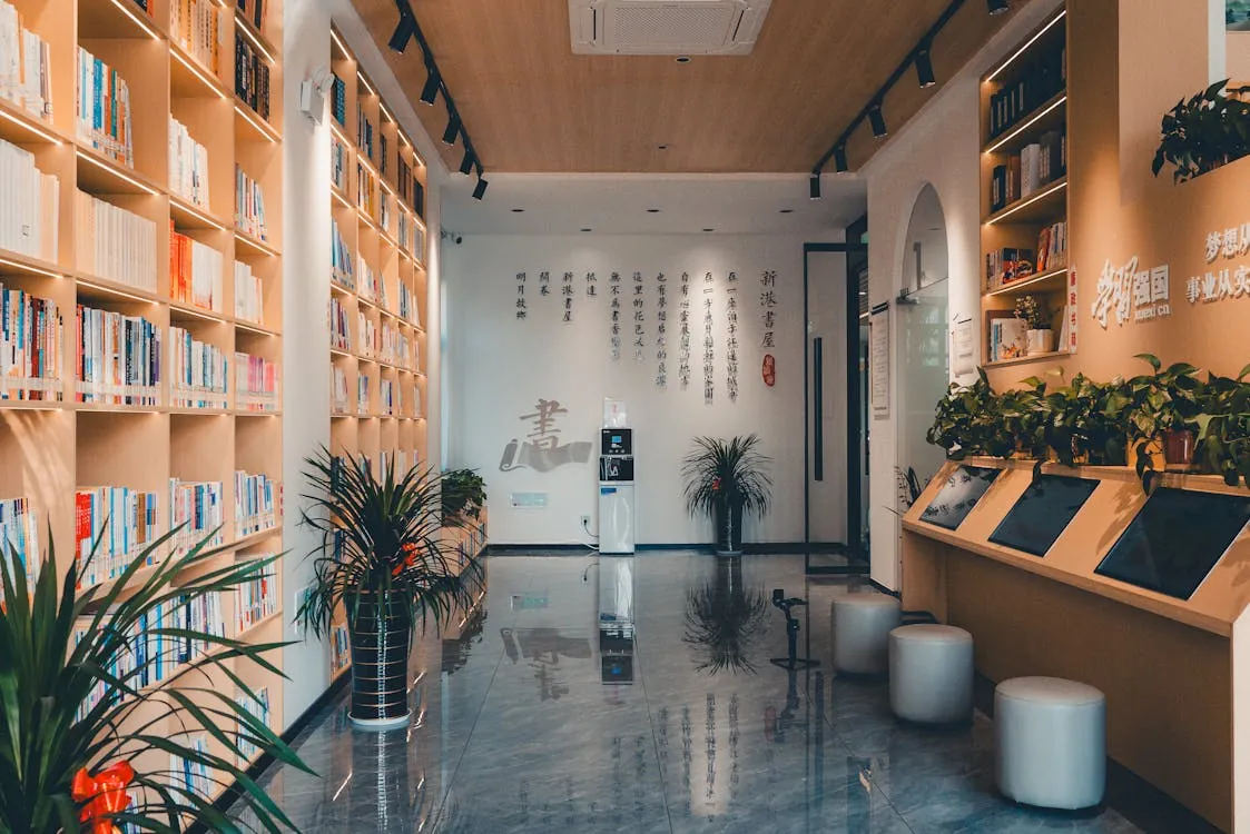 This image depicts a modern, well-lit library or reading area with floor-to-ceiling bookshelves filled with colorful books on the left, indoor plants adding greenery, and a wall featuring Chinese calligraphy or artwork in the background; the space also includes tablet stations on the right and seating options, creating a cozy and inviting environment suitable for reading or study purposes.
