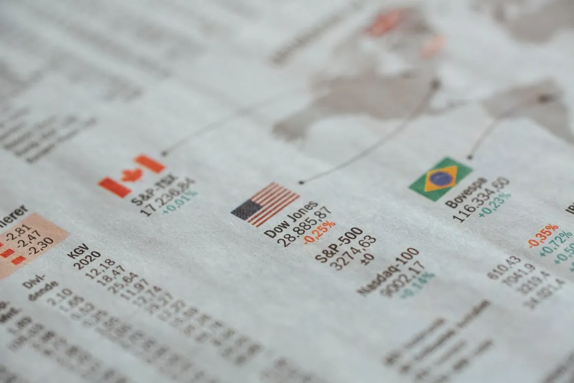 This close-up image shows a financial newspaper featuring stock market data, including the Dow Jones, S&P 500, and Nasdaq indices, with notable figures highlighted in red and green to indicate market declines and gains. The setting appears to be a typical financial news page, suitable for articles about stock performance, economic analysis, or investment updates.
