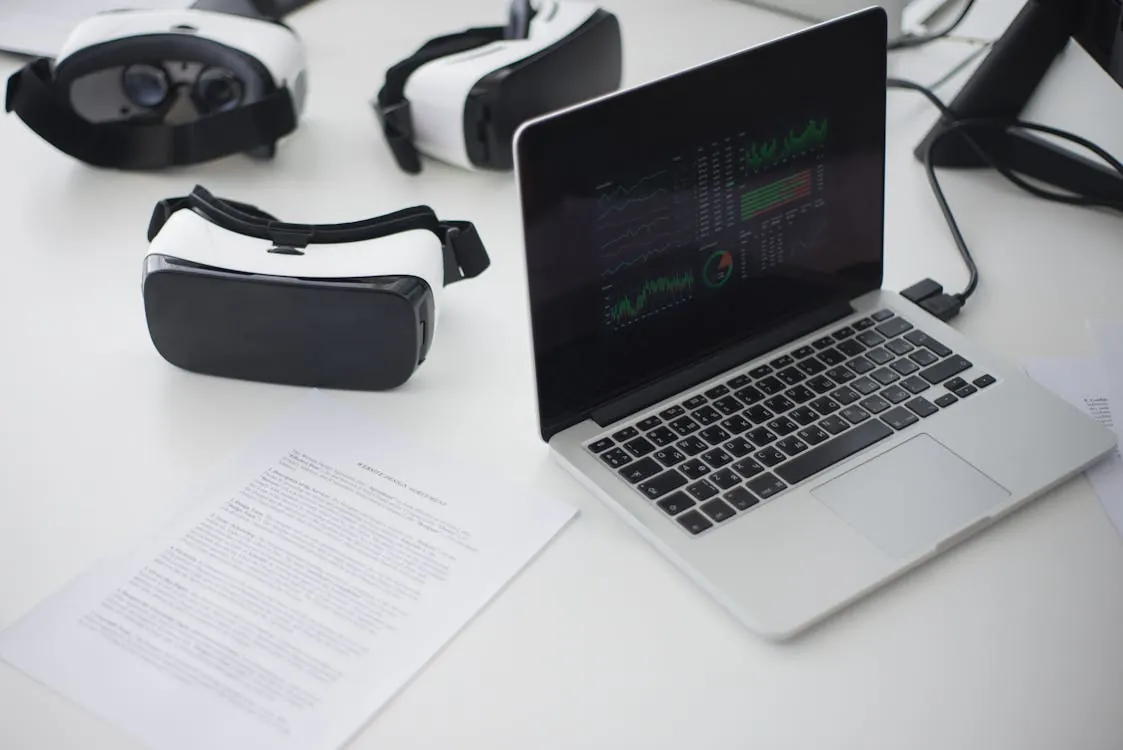 The image shows a workspace featuring two virtual reality headsets, a laptop displaying data visualizations, and printed documents on a white desk. It would suit an article about virtual reality technology, digital data analysis, or the integration of VR in professional settings.