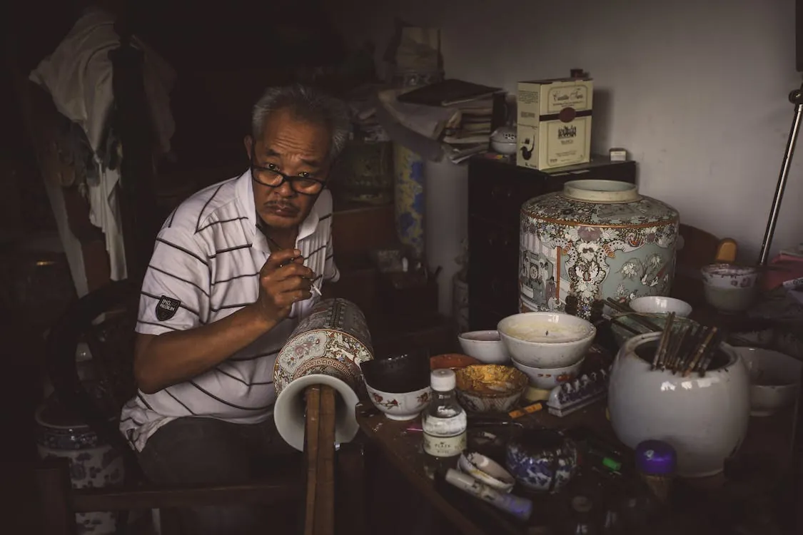 An elderly craftsman meticulously paints a porcelain vase with detailed patterns in his workshop.