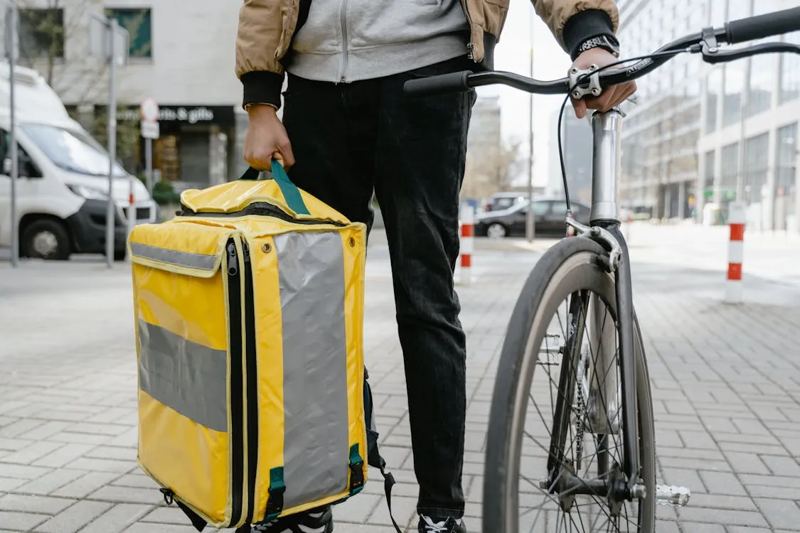 Courier with insulated delivery backpack holding a bicycle in a city setting