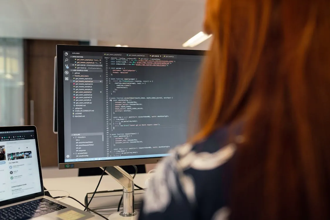 The photo shows a person with long reddish hair working on a programming project, focusing on a computer monitor displaying code, with a laptop open beside it displaying a social media profile. The setting appears to be a modern office or workspace, highlighting software development or coding activities.