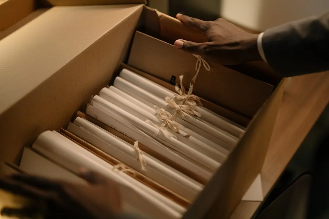 The image shows a person in a suit reaching into a cardboard box filled with neatly organized rolled-up documents or certificates tied with beige ribbons, suggesting a professional or formal setting, possibly related to an event like a wedding, graduation, or awards ceremony. The warm lighting and careful arrangement emphasize organization, presentation, or special significance of the items.
