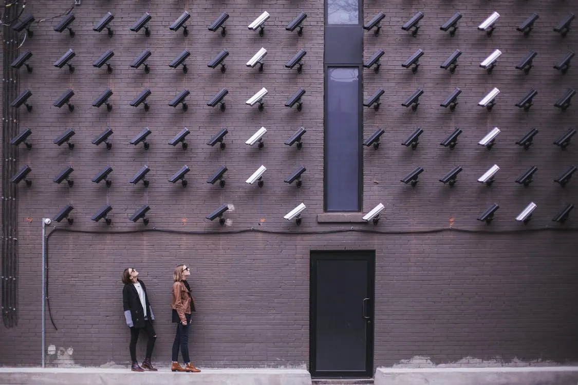 This photo shows two women standing and looking up at a modern building's brick wall, which is decorated with an array of surveillance cameras arranged in a pattern. The setting emphasizes themes of security, privacy, or urban surveillance technology.