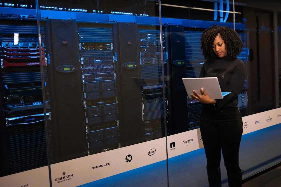 The image shows a woman standing in front of a row of server racks in a data center, holding a laptop and examining the equipment. Notable details include the blue lighting and visible brand logos on the server cabinets, suggesting a high-tech environment suitable for discussions about data infrastructure, cybersecurity, or digital technology.