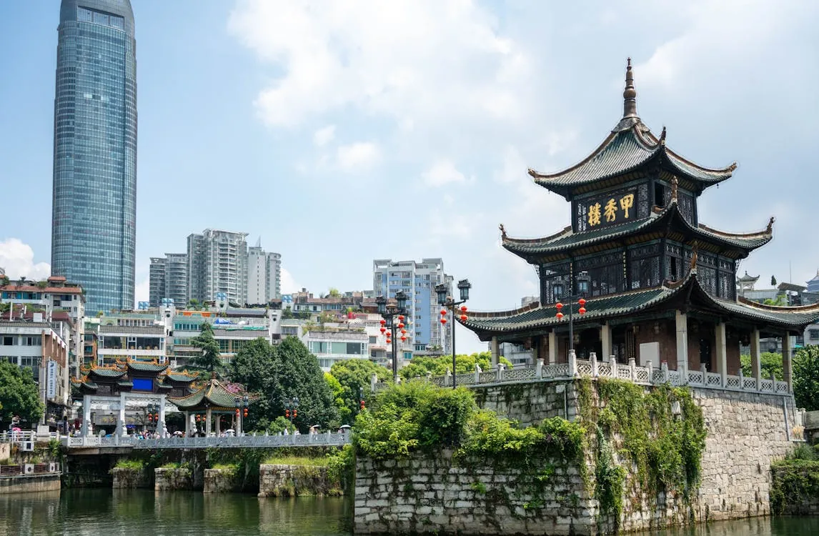 This photo shows a traditional Chinese-style pavilion situated on a small stone island with a bridge, set against a modern city skyline with tall buildings, including a prominent glass skyscraper, under a partly cloudy sky. The scene combines historic architecture with contemporary urban development, suitable for articles about cultural heritage amidst modernization.