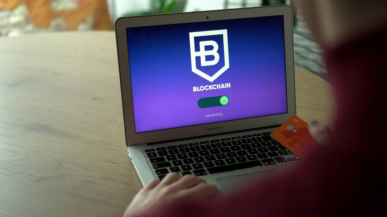 The image shows a person holding a credit card while sitting in front of a MacBook Air, which displays a screen with a blockchain logo and the text "Connecting..." indicating an attempt to access or set up a blockchain-based service or wallet. This setting suggests a focus on digital finance, cryptocurrency, or blockchain technology in a casual workspace environment.