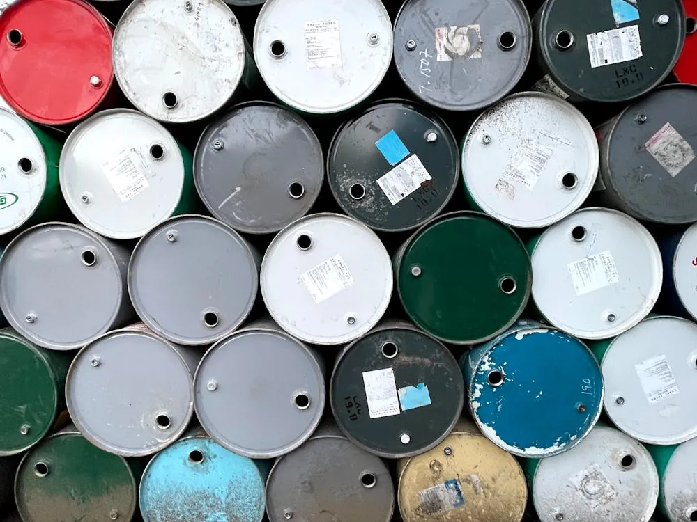 This image shows a close-up view of multiple industrial metal drums or barrels stacked together, featuring various colors such as red, white, black, blue, green, and gray. The barrels have visible labels, some with barcodes and wear marks, indicating they are used for storage or transportation of chemicals or liquids.