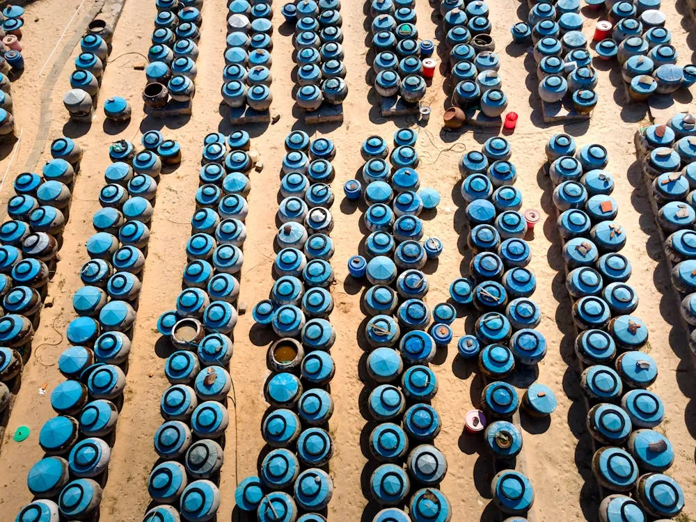 This aerial photo shows numerous plastic buckets with blue covers arranged in neat, parallel rows on a sandy surface, likely at a construction site, outdoor market, or storage area. The uniformity and organization suggest they are prepared for a large-scale activity or distribution.