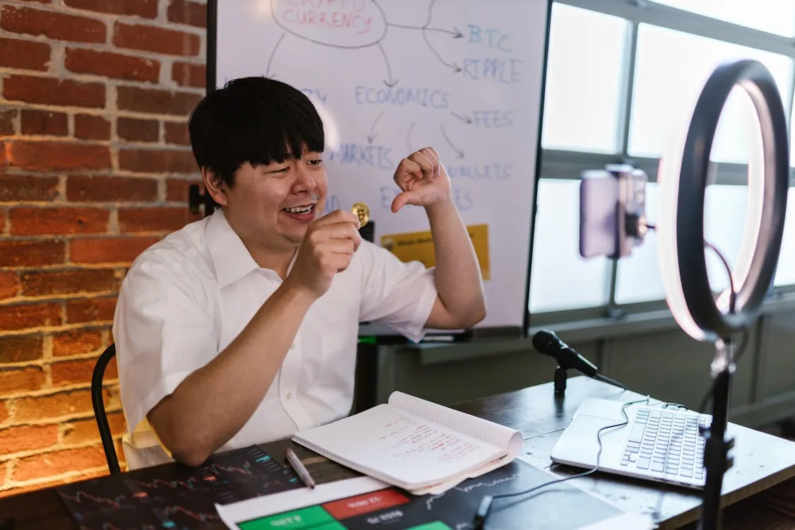 This photo shows a man sitting at a desk in a modern office setting, celebrating as he holds a gold coin. The workspace includes a laptop, a notebook with handwritten notes, a microphone, and a ring light, with a whiteboard in the background featuring financial and cryptocurrency-related diagrams.