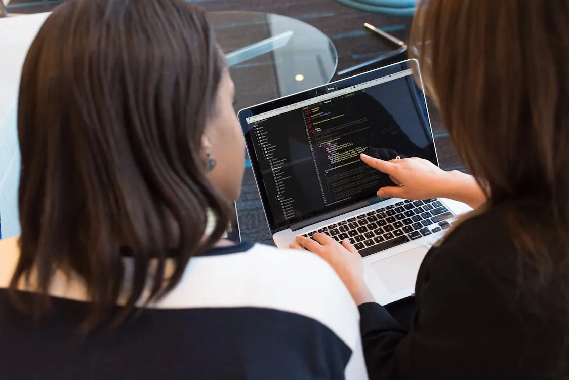 The photo shows two women collaborating over a laptop displaying lines of code, suggesting a programming or software development setting, likely in an office or conference room. This image would be suitable for articles about teamwork in tech, coding projects, or women in technology.