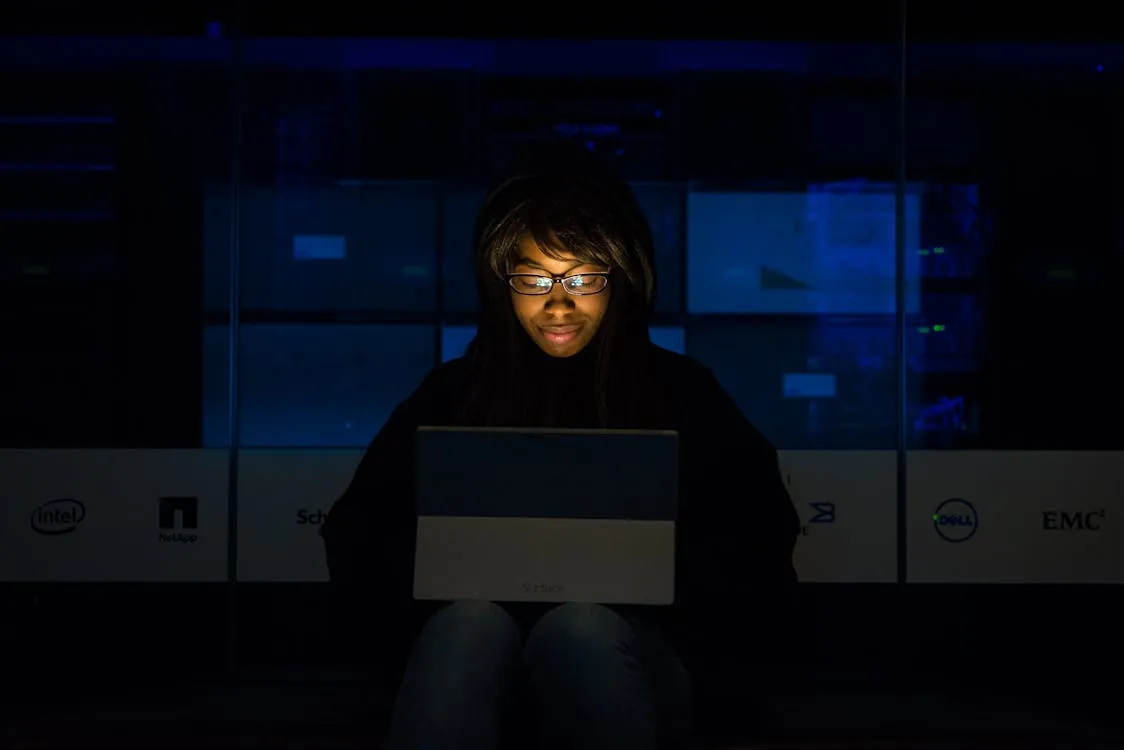 This image shows a young woman with glasses working on a laptop in a dark environment, likely a data center or technology lab, with only her face and device illuminated. Notable details include the glowing screen and the logos of technology companies in the background, indicating a focus on tech or cybersecurity themes.