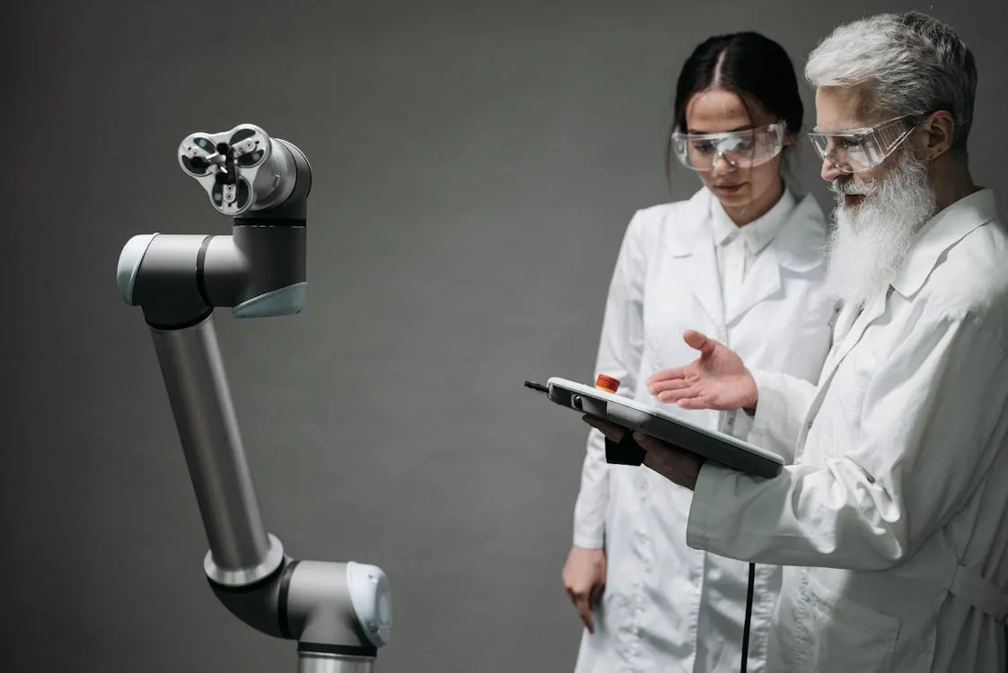 The image shows two scientists, a man with a long white beard and a woman, both wearing white lab coats and safety goggles, examining a robotic arm in a laboratory setting. The scene emphasizes robotics and advanced technology, suitable for articles on scientific innovation, robotics development, or future technological advancements.