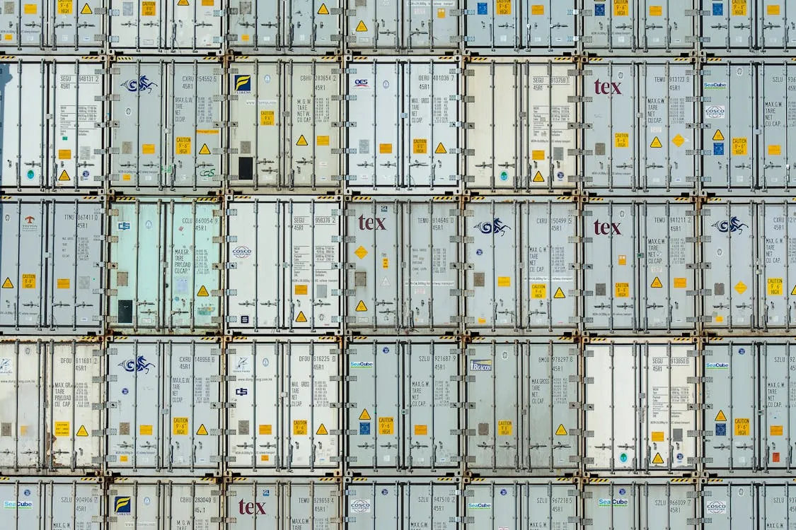 This photo shows a stacked arrangement of shipping containers in a warehouse or port setting, with a focus on their uniform design, safety markings, and branding labels, suggesting themes of logistics, trade, or transportation. The image could complement articles discussing global supply chains, shipping industries, or infrastructure.
