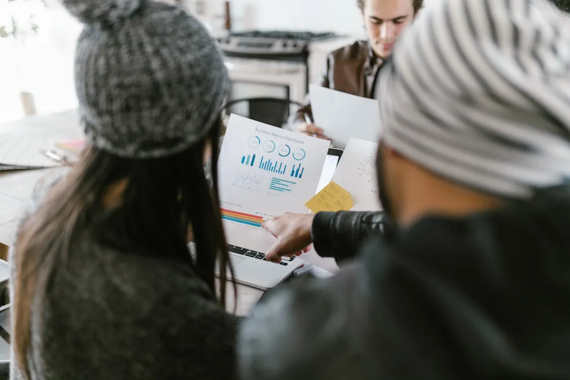 The photo shows a group of three young professionals analyzing business data and charts on a laptop and printed reports in a modern office setting, with a focus on collaboration and data sharing. This image would suit articles about teamwork, business analytics, or startup meetings.