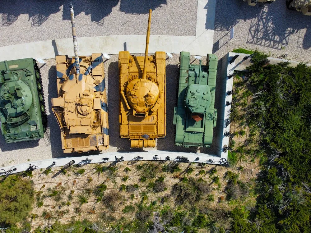 This aerial photo shows four tanks of different colors (green, tan, yellow, and green) displayed outdoors on a paved area with a surrounding white wall, next to a wooded area. The image highlights a military or museum exhibit setting, suitable for articles about military history, vehicle collections, or outdoor displays.
