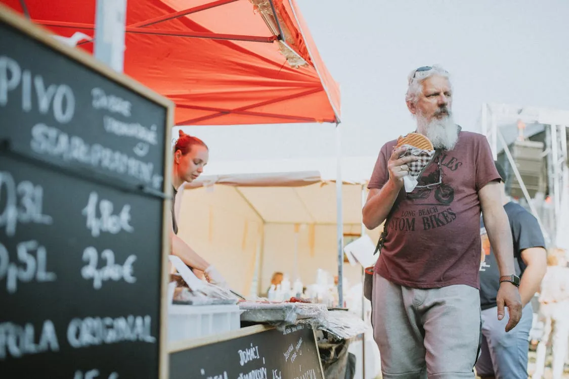 This image shows a man with a long white beard and casual clothing holding a sandwich at an outdoor market or food fair, with a vendor in the background preparing food under a red canopy. The setting appears lively and casual, suitable for an article about food festivals, outdoor markets, or diverse food experiences.