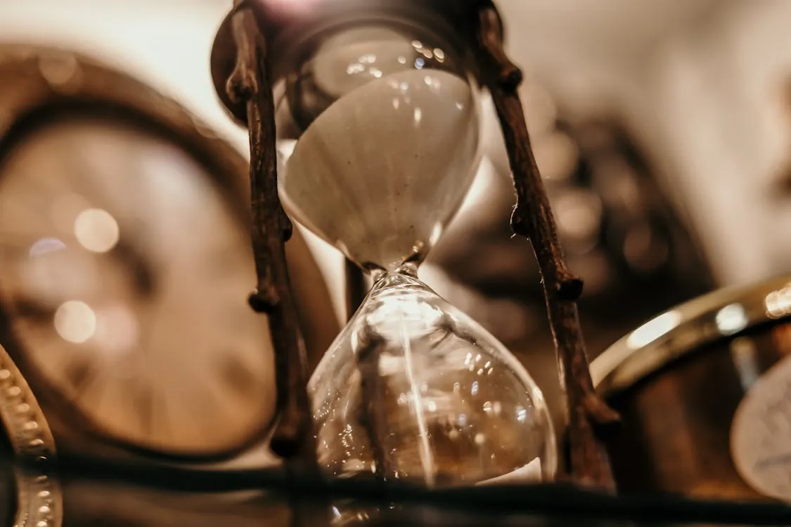 This close-up image features an hourglass with white sand, encased in a rustic metal frame, surrounded by multiple clocks, suggesting themes of time, aging, or deadlines. It would be suitable for articles related to time management, the passage of time, or vintage/scenic decoration.