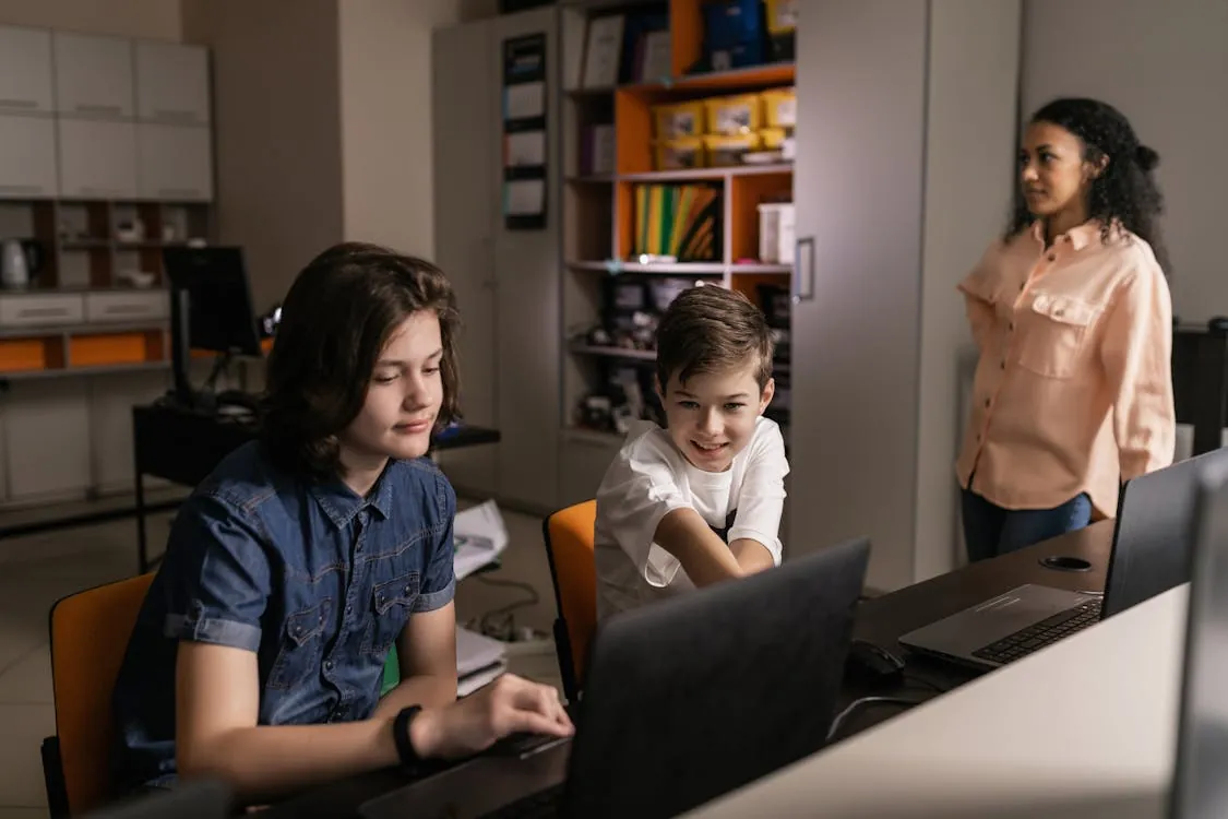 The photo shows three children in a classroom or study environment, with two boys seated at desks using laptops and a girl standing nearby, all engaged in a digital learning activity. The setting features shelves with books and educational materials, highlighting a focus on technology-enabled education or collaborative learning.