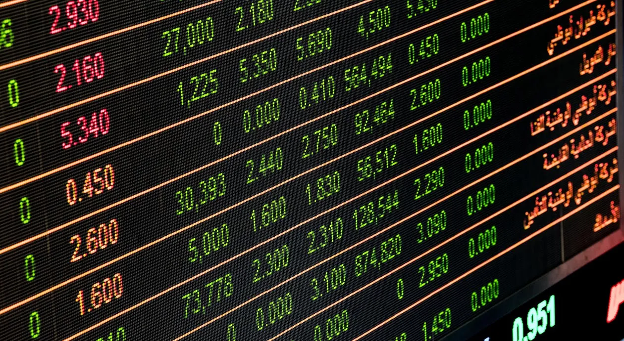 This photo shows a digital stock market display board with rows of stock prices and trading volumes, featuring bright green and red text indicating gains and losses, set against a dark background. It captures the dynamic and high-tech atmosphere of financial trading environments.