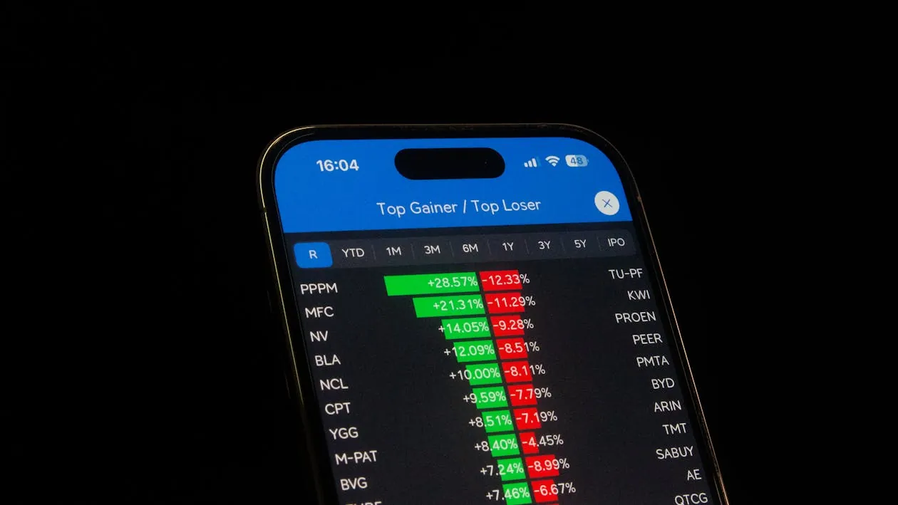 This photo shows a smartphone screen displaying a financial stock market app focused on "Top Gainer/Top Loser" stocks, with green and red percentages indicating stock performance over a specified period. The setting is dark, highlighting the illuminated screen, making it suitable for articles about investing, stock analysis, or financial technology.