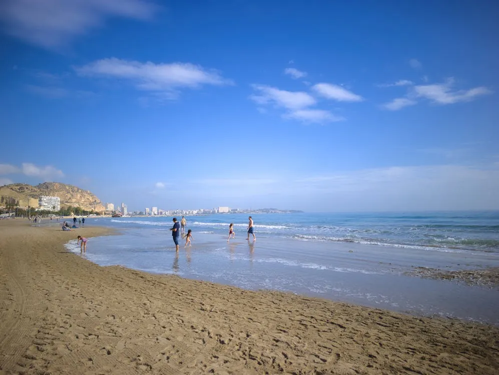 The photo shows a sandy beach with several families and children playing near the shoreline under a bright blue sky with some clouds, with a cityscape and hills visible in the background, suggesting a coastal urban setting ideal for articles about beach destinations or family travel.