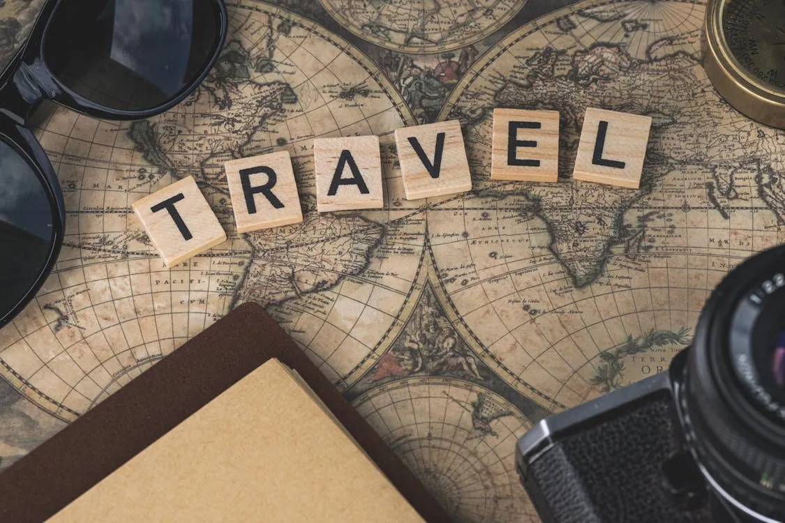 This photo features a vintage world map as the background with the word "TRAVEL" spelled out in wooden letter tiles in the center. It also includes travel essentials like sunglasses, a camera, and a notebook, evoking a theme of adventure and exploration.