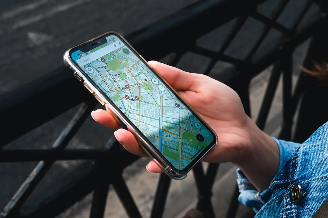 The image shows a person holding a smartphone displaying a detailed city map with navigation routes, set outdoors on a bench or table with a dark, blurred background and metal railing. This photo emphasizes mobile navigation or GPS technology, suitable for articles about travel, apps, or urban exploration.