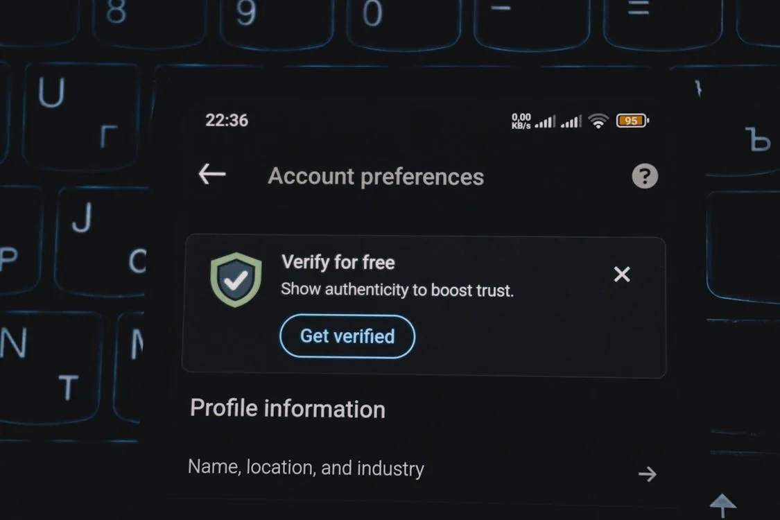 The photo shows a smartphone screen displaying an "Account preferences" page with a prominent option to verify the account for free, including a "Get verified" button. It appears to be taken in a dimly lit environment, possibly at night, with a keyboard partially visible behind the phone.