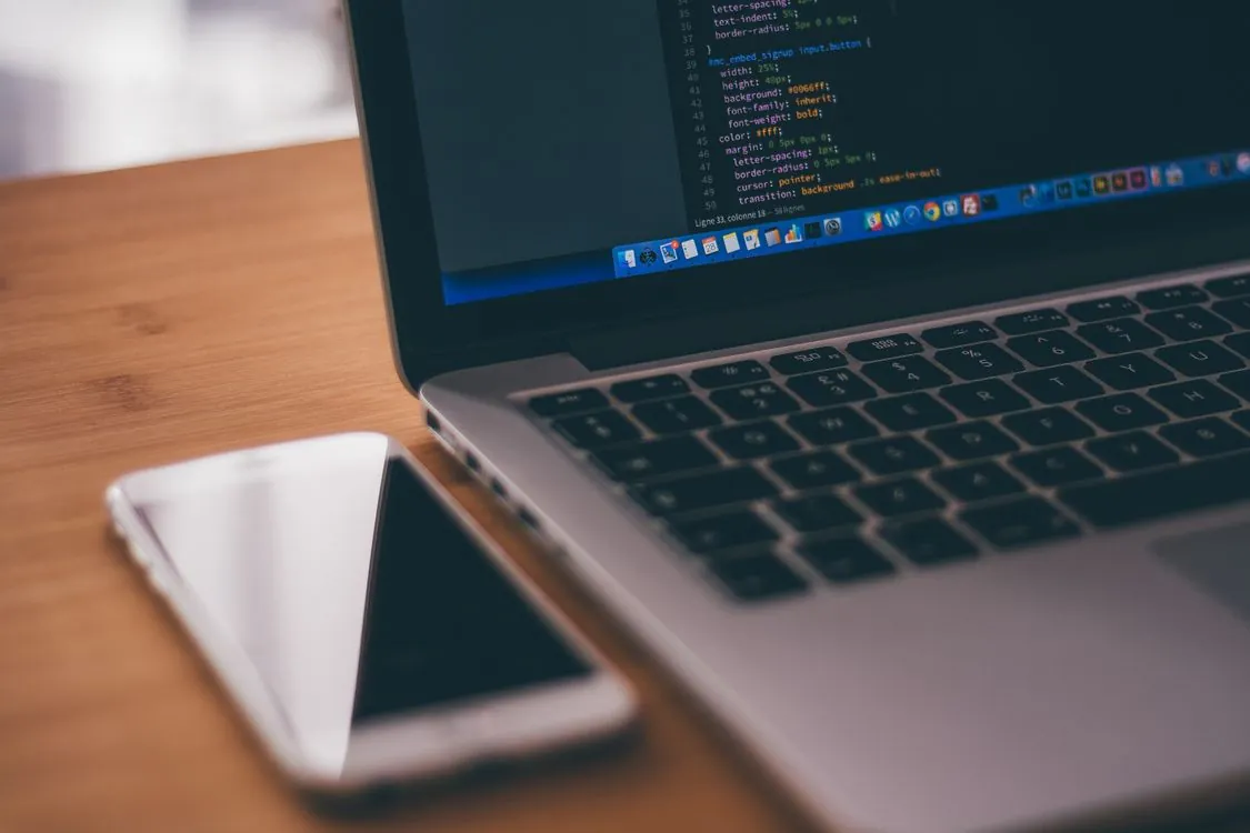 Mobile app developer writing code on laptop with smartphone on desk